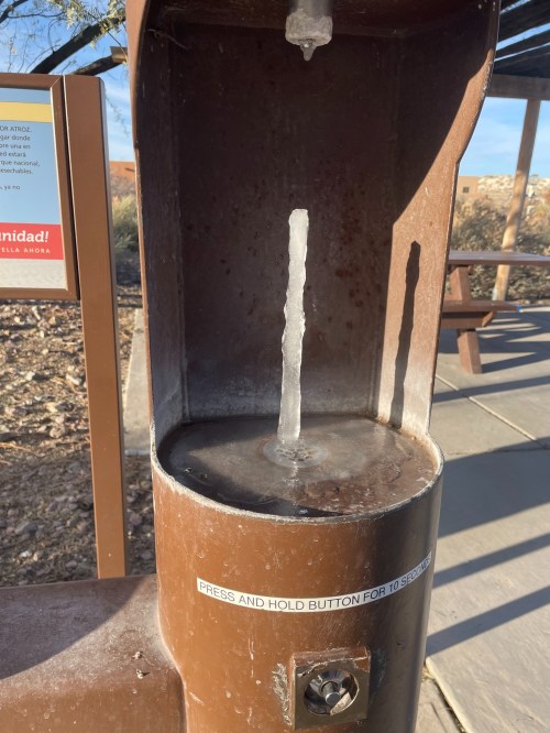 A water fountain with a frozen stream of water, surrounded by a desert landscape and a sign nearby.