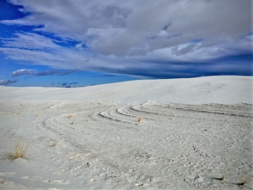 A vast white landscape with gentle sand dunes under a cloudy blue sky.