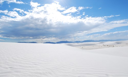 A vast white sand dune landscape under a bright blue sky with scattered clouds and distant mountains.