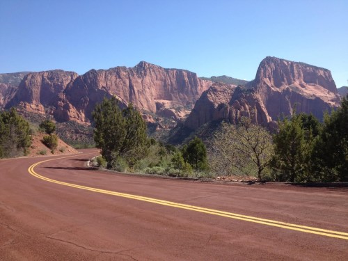 A winding road curves through a scenic landscape of red rock mountains and clear blue skies.