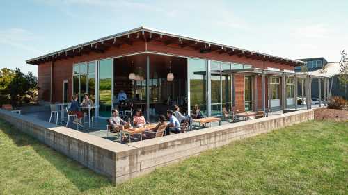 A modern building with large windows, surrounded by grass, where people are socializing and working outside.