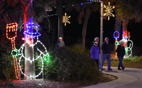 Colorful holiday lights decorate trees, featuring snowmen and stars, as people stroll along a path at night.