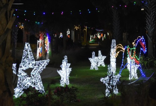 Colorful holiday lights illuminate a park, featuring star and horse decorations amidst greenery.