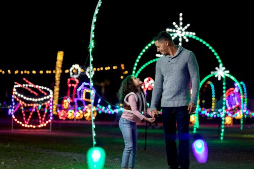 A man and a girl hold hands, smiling at each other, surrounded by colorful holiday lights at night.