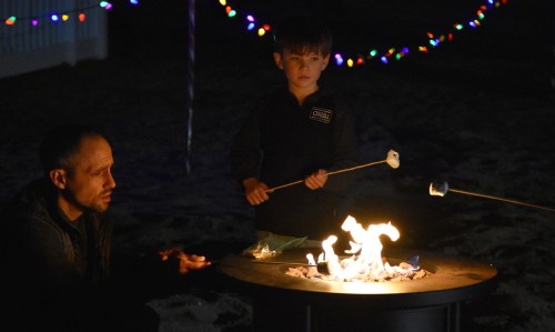A man and a boy roast marshmallows over a fire pit, surrounded by colorful string lights at night.