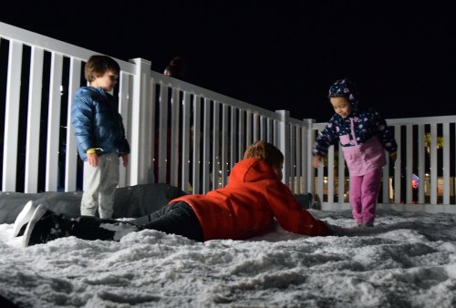 Two children play in the snow at night while a third child lies on the ground, surrounded by a white fence.