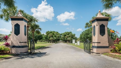 A scenic entrance with decorative gates and palm trees, leading to a lush green pathway under a blue sky.