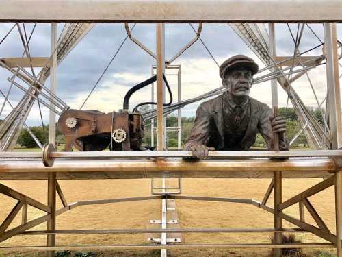 A bronze statue of a man in a cap, leaning on a railing, with a vintage machine beside him, set against a cloudy sky.