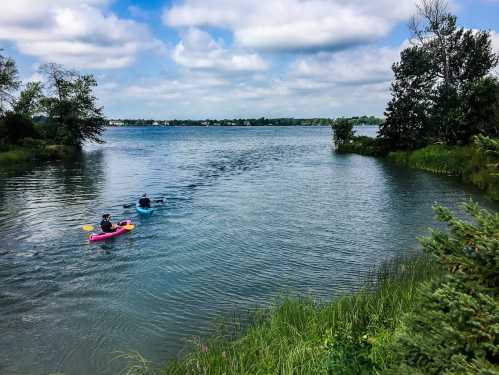 Two people kayaking on a calm lake surrounded by greenery and cloudy skies.