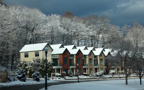 Row of houses with snow-covered roofs, surrounded by trees with frost, under a cloudy sky.