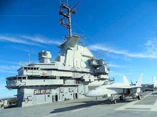 A military aircraft carrier with a fighter jet on the deck, under a clear blue sky.
