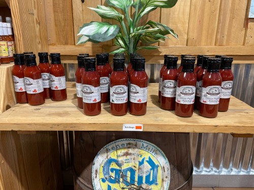 A wooden shelf displaying multiple bottles of ketchup, with a plant in the background and a decorative barrel below.