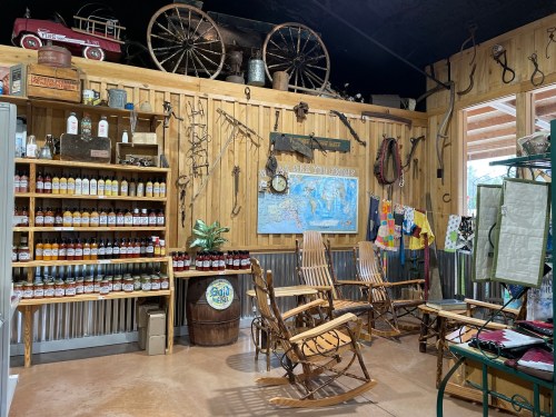 A rustic interior featuring a wooden wall, shelves of sauces, rocking chairs, and vintage decor items.
