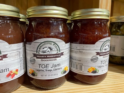 Two jars of "Toe Jam" jam on a wooden shelf, labeled with flavors: tangerine, orange, and elderberry.