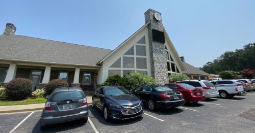 A building with a stone facade and clock tower, surrounded by parked cars on a sunny day.