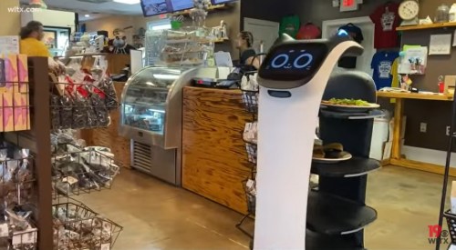 A robot waiter stands in a café, surrounded by customers and a display of baked goods and snacks.