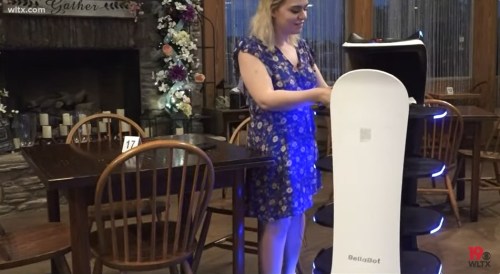 A woman interacts with a robotic server in a restaurant, with a cozy fireplace and wooden tables in the background.