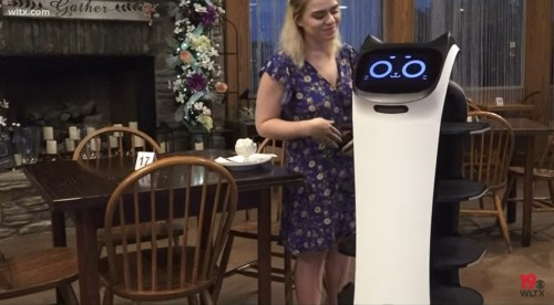 A woman in a floral dress interacts with a friendly robot waiter in a cozy restaurant setting.