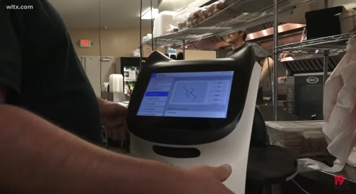 A person interacts with a touchscreen robot in a kitchen setting, with shelves and food items in the background.