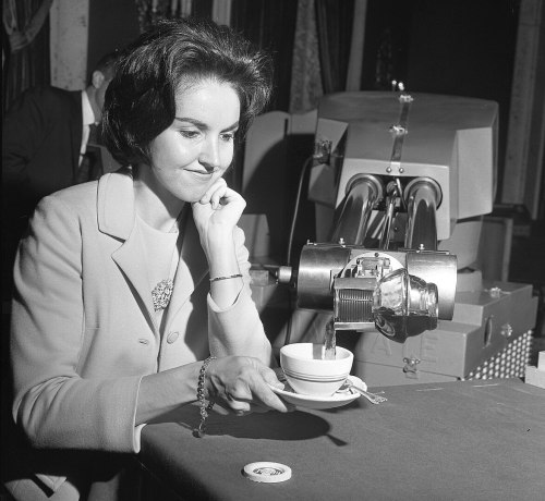 A woman in a light-colored suit sits at a table, pouring coffee from a machine into a cup.