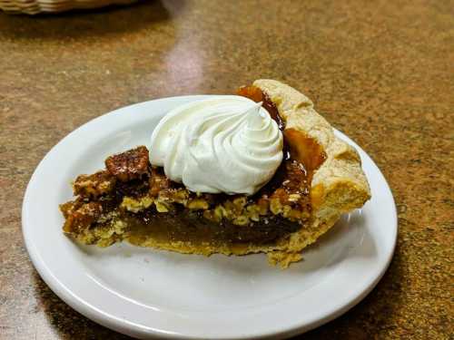 A slice of pecan pie topped with whipped cream on a white plate, resting on a wooden table.