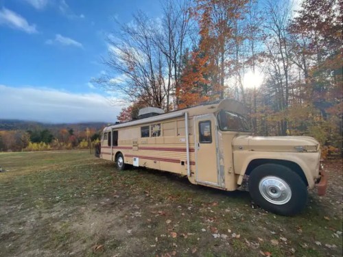 A vintage bus parked in a scenic area with autumn trees and a cloudy sky in the background.