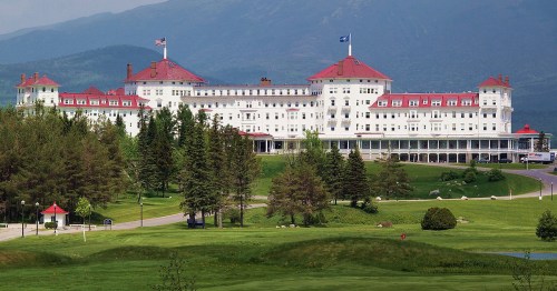 A grand white hotel with red roofs, surrounded by green trees and mountains in the background.