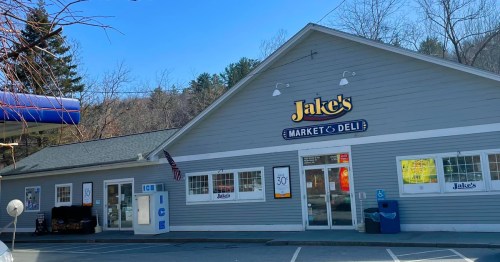 Exterior of Jake's Market & Deli, featuring a gray building with signage and a parking area. Trees in the background.