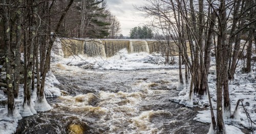 A snowy landscape featuring a waterfall cascading over rocks, surrounded by trees and a rushing river below.
