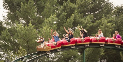 A roller coaster with excited riders raising their hands, surrounded by tall green trees.