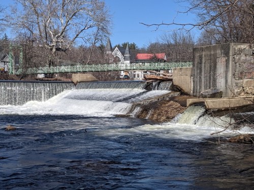 A flowing river over a dam with a green bridge in the background and trees lining the shore under a clear blue sky.