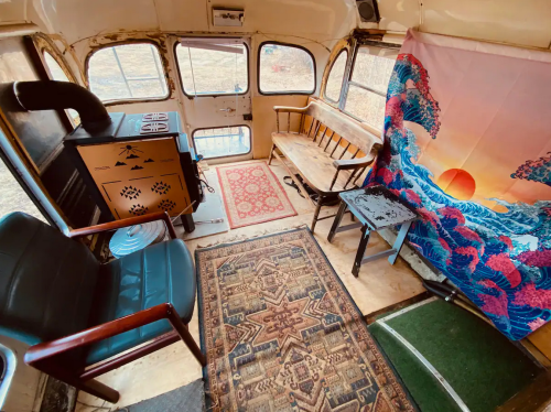 Interior of a converted bus featuring a rug, wooden benches, a small table, and a colorful wall hanging.