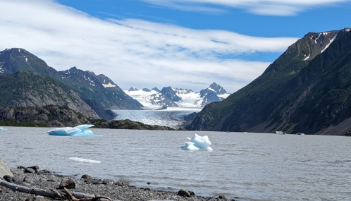 A serene landscape featuring a glacial lake, icebergs, and mountains under a blue sky with scattered clouds.