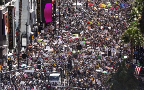 A large crowd of protesters fills a city street, holding signs and banners during a demonstration.