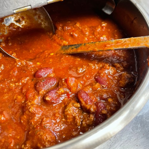 A close-up of a pot of chili with beans and meat, featuring a metal ladle and wooden spoon.