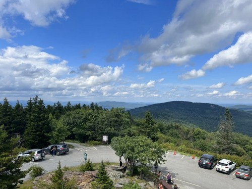 A scenic view from a mountain overlook, featuring a parking area, lush trees, and a blue sky with clouds.