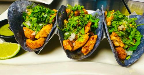 Three blue corn tacos filled with grilled chicken, topped with cilantro and onions, served with a lime wedge and green salsa.