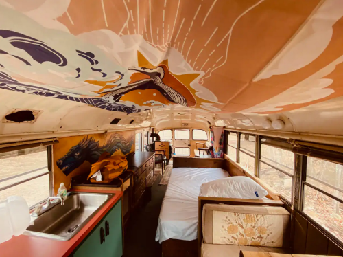 Interior of a converted bus featuring a bed, kitchen area, and colorful ceiling mural of a whale and sun.