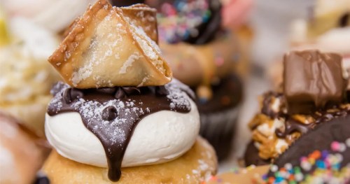 A close-up of a cupcake topped with chocolate sauce, whipped cream, and a pastry triangle, surrounded by colorful desserts.
