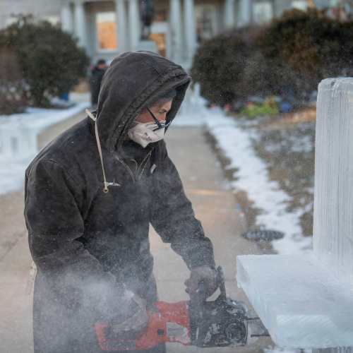 A person in a mask and hoodie uses a tool to carve or shape a block of ice outdoors in a snowy environment.