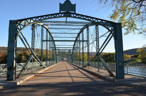A green metal bridge spans a river, with a clear blue sky and trees in the background, showcasing a peaceful landscape.