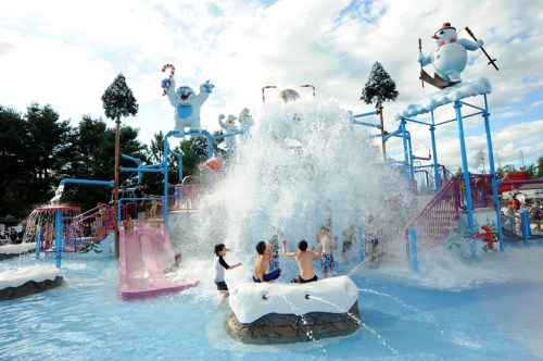 Children play in a water park with slides and a large splash feature, surrounded by festive decorations and trees.