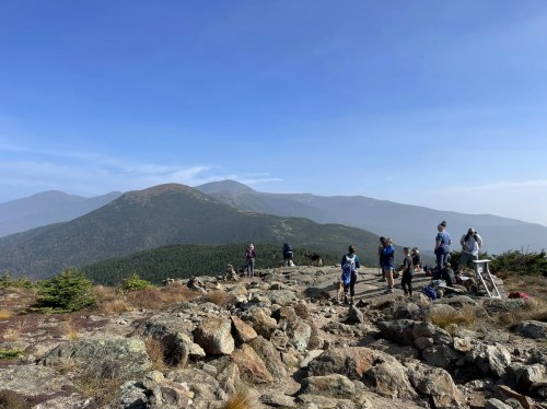 Hikers on rocky terrain overlooking a mountain range under a clear blue sky.