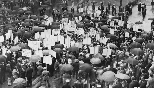A large crowd with umbrellas holding protest signs in a rainy urban setting, advocating for various social issues.