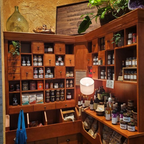 A wooden display filled with jars and bottles of various spices and herbs, with plants and a lamp in a cozy shop setting.