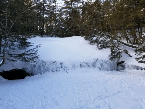 A snowy landscape with a frozen waterfall and trees in the background, creating a serene winter scene.