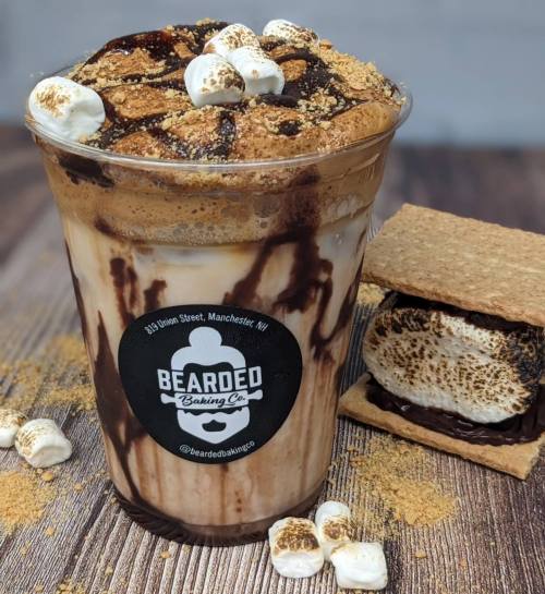 A delicious layered drink topped with marshmallows and graham cracker crumbs, next to a s'more on a wooden surface.