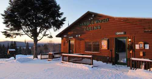 A wooden cabin with "Ramblewood Cabins" sign, surrounded by snow and trees, during sunset.