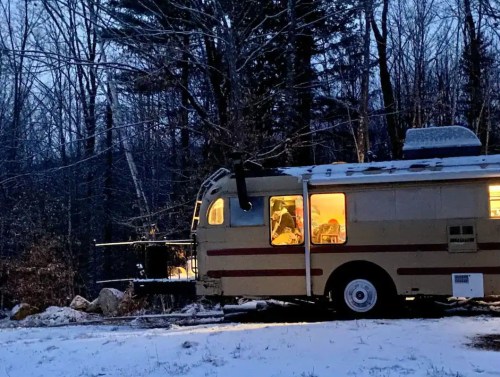 A vintage camper parked in a snowy forest, with warm light glowing from its windows at dusk.