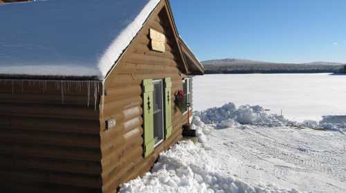 A cozy log cabin with green shutters, surrounded by snow, overlooking a frozen lake under a clear blue sky.